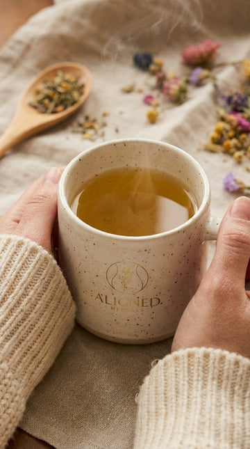 Hands wrapped around an Aligned Herbals branded ceramic mug filled with steaming herbal tea surrounded by dried wildflowers and a wooden spoon