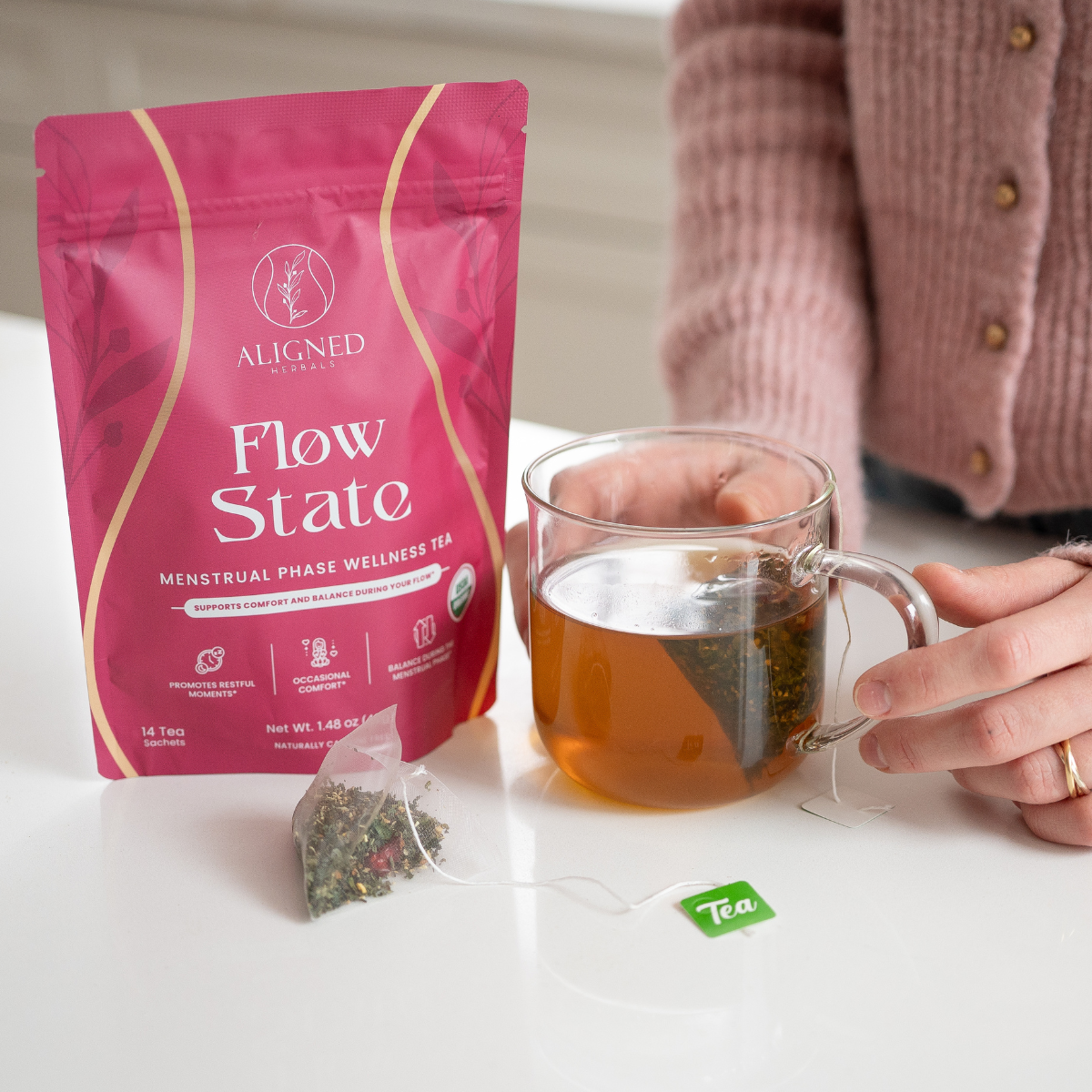 Aligned Herbals Flow State menstrual phase wellness tea bag next to a glass mug of steeping herbal tea held by a woman in a pink cardigan