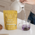 Hand pouring hot water from a white kettle into a glass mug of Aligned Herbals Peak Power ovulation phase tea turning purple next to the yellow product bag