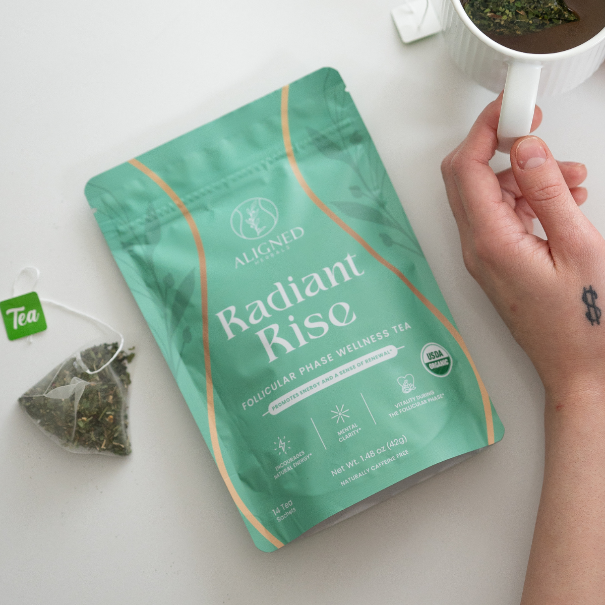 Aligned Herbals Radiant Rise follicular phase wellness tea bag laying flat next to a pyramid tea sachet and a hand holding a white ceramic mug