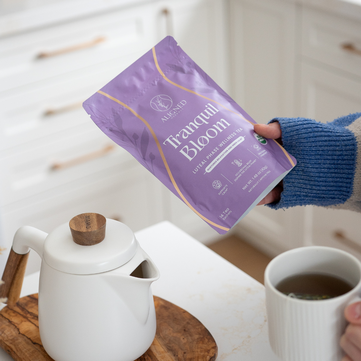 Hand holding Aligned Herbals Tranquil Bloom luteal phase wellness tea bag above a white teapot and ceramic mug of herbal tea in a bright kitchen