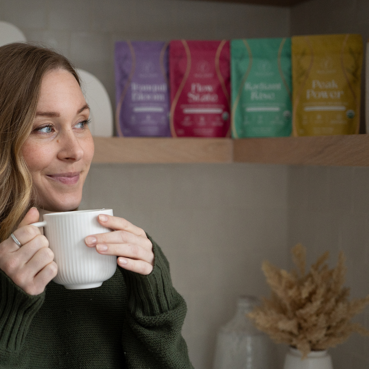 Woman in a green sweater holding a white ribbed mug with all four Aligned Herbals cycle syncing tea bags displayed on a wooden shelf behind her